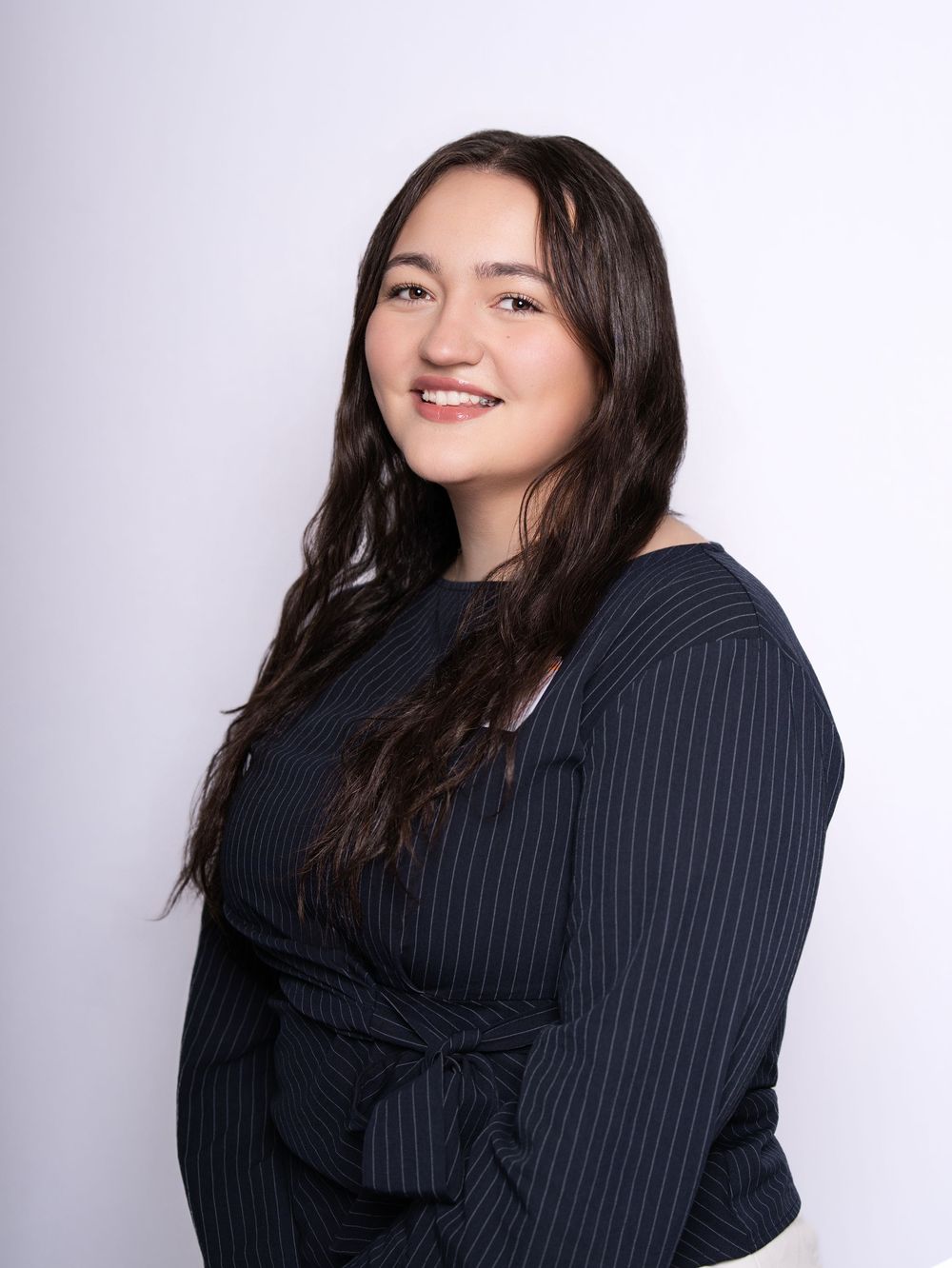 A smiling woman with long dark hair in a navy pinstripe top against a light studio backdrop