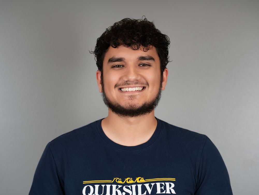 A smiling young man with curly hair and a beard in a navy T-shirt against a grey backdrop