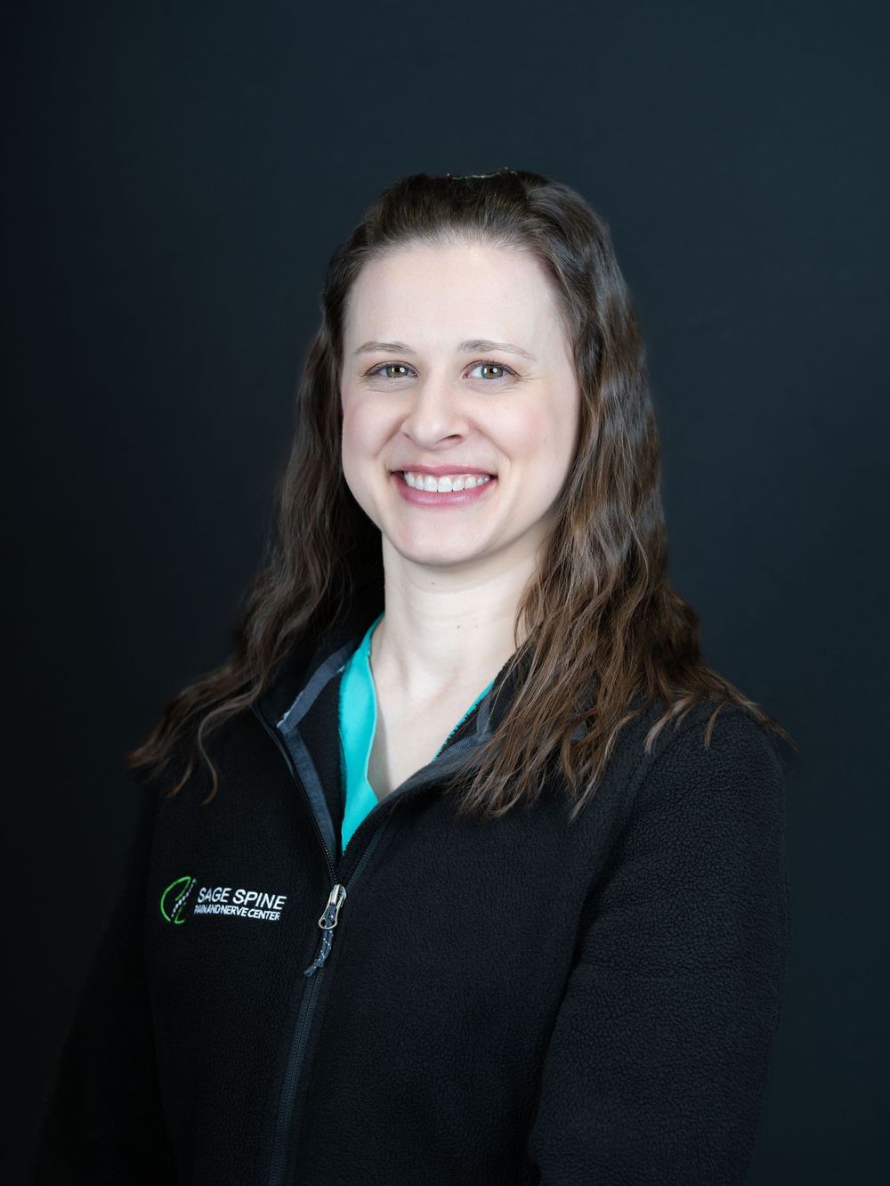 A smiling woman with wavy brown hair in a black zip-up jacket against a dark studio backdrop