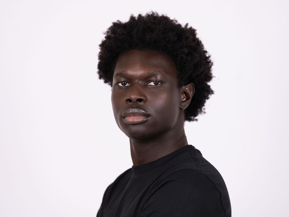 A young man in a black top with a serious expression against a light studio backdrop