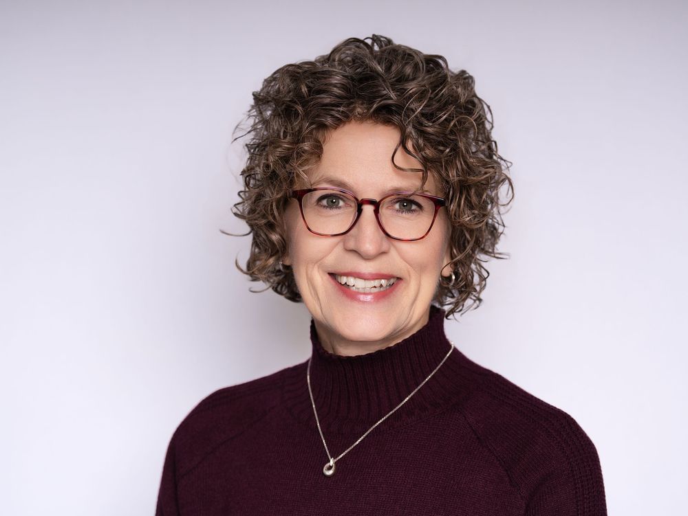 A smiling woman with curly hair and red glasses in a burgundy turtleneck against a light backdrop