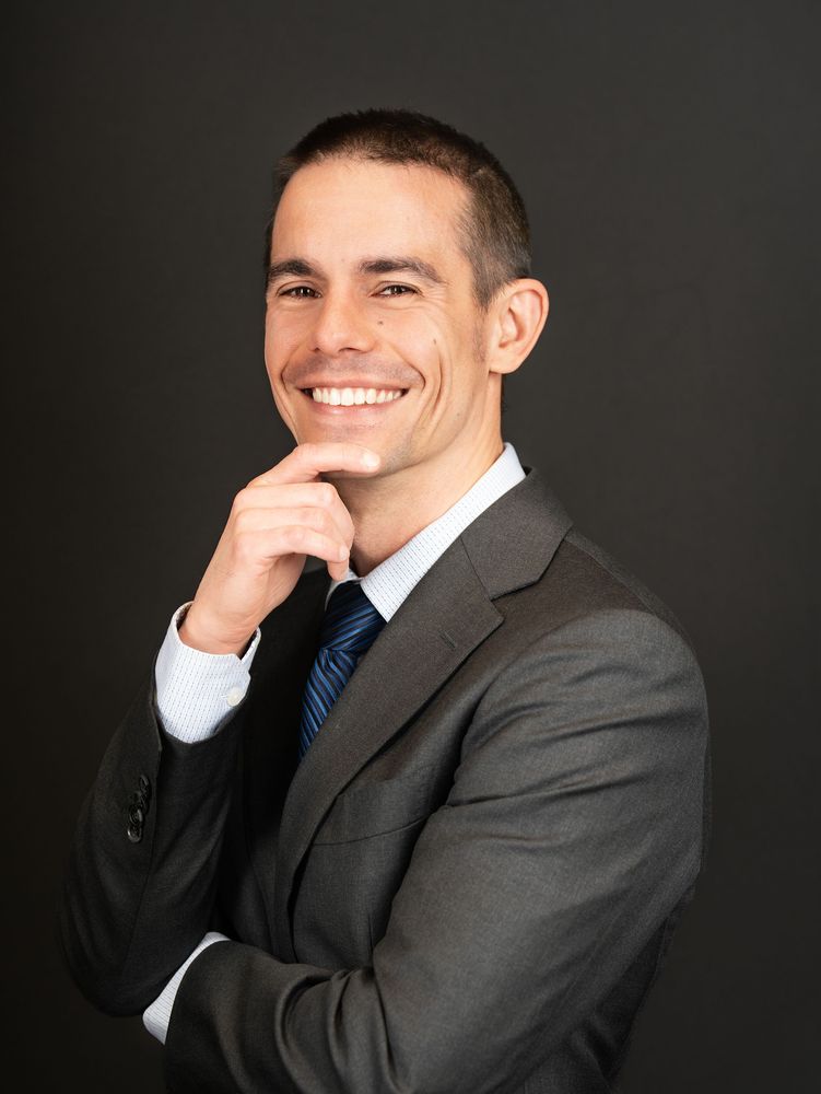 A smiling man in a grey suit and tie with his hand at his chin, dark backdrop