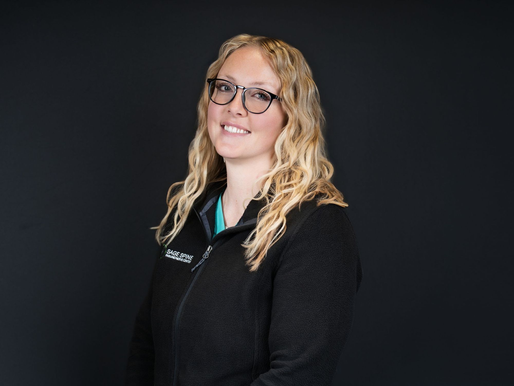 A smiling woman with curly blonde hair and glasses in a black zip-up jacket against a dark backdrop