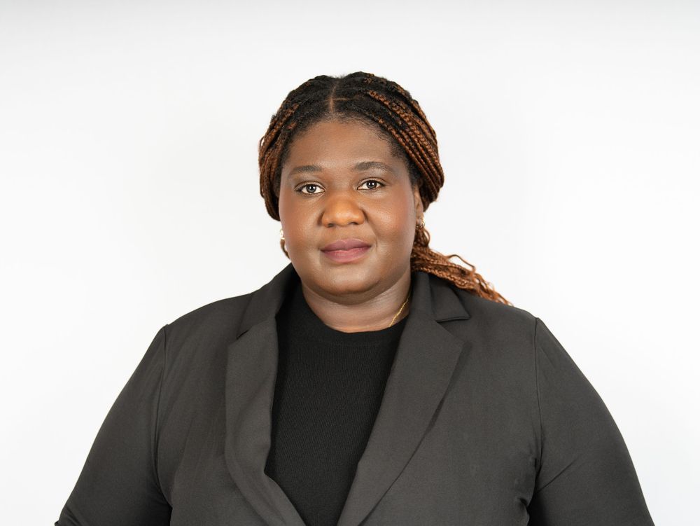A woman with long braids in a charcoal blazer against a white studio backdrop