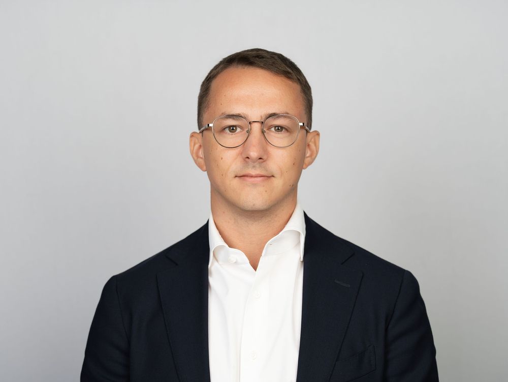 A man with glasses in a navy blazer and white shirt against a grey studio backdrop