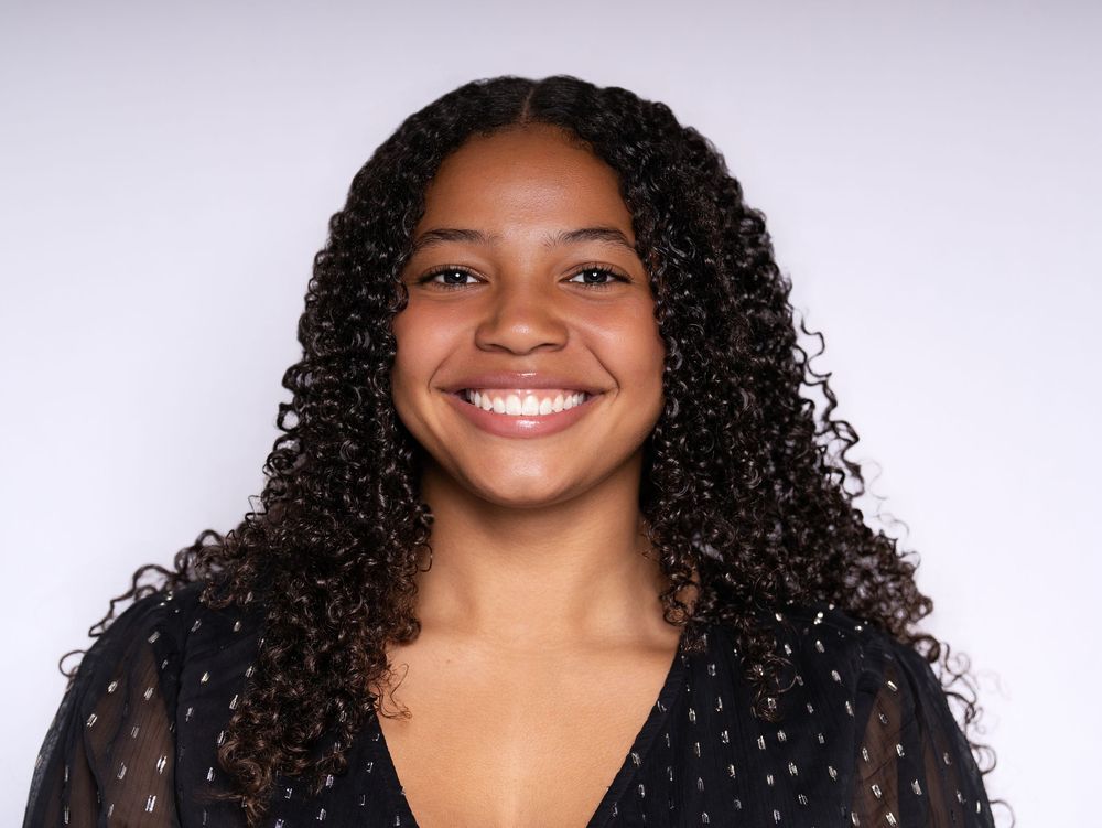 A smiling young woman with long curly hair in a black top against a light studio backdrop