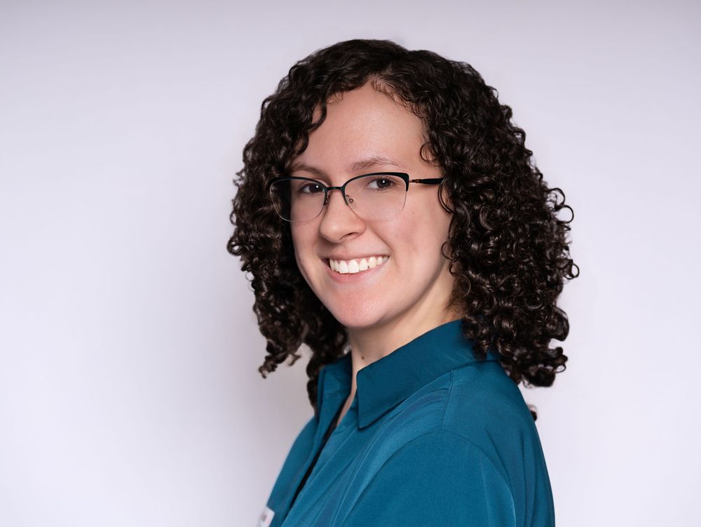A smiling woman with curly hair and glasses in a teal blouse looking over her shoulder