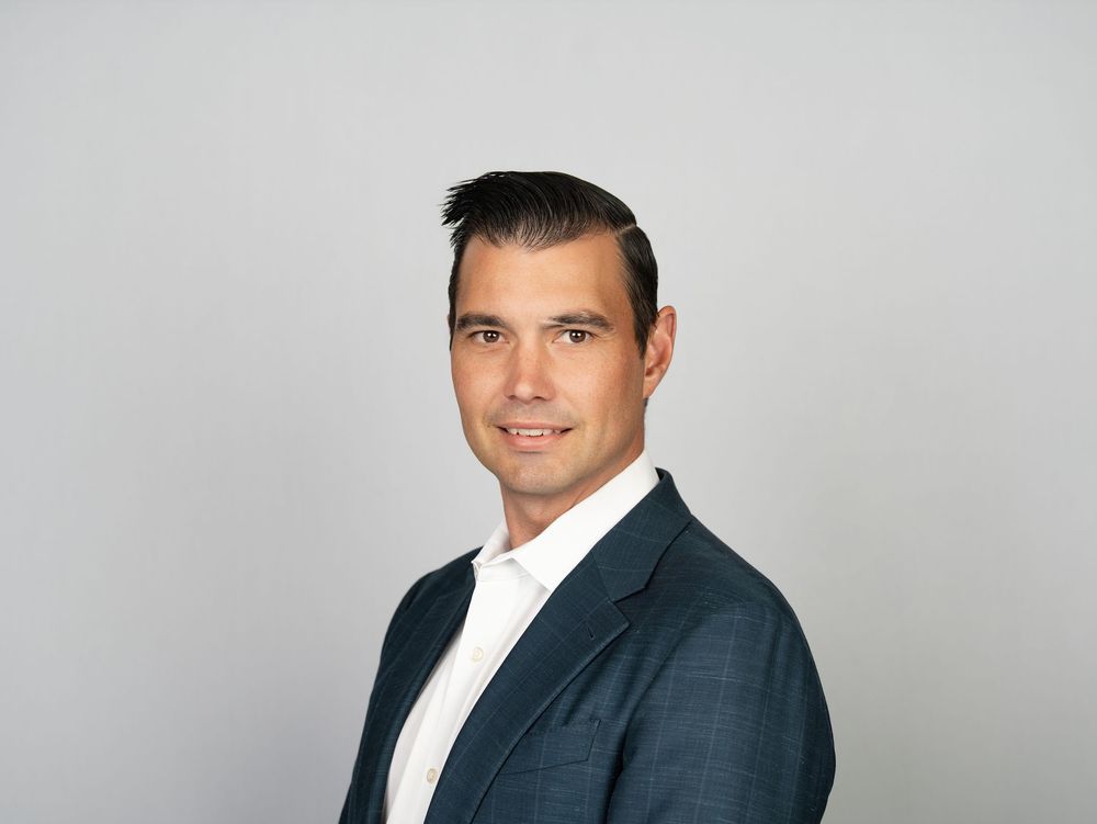 A man in a navy blazer and open-collar white shirt against a grey studio backdrop