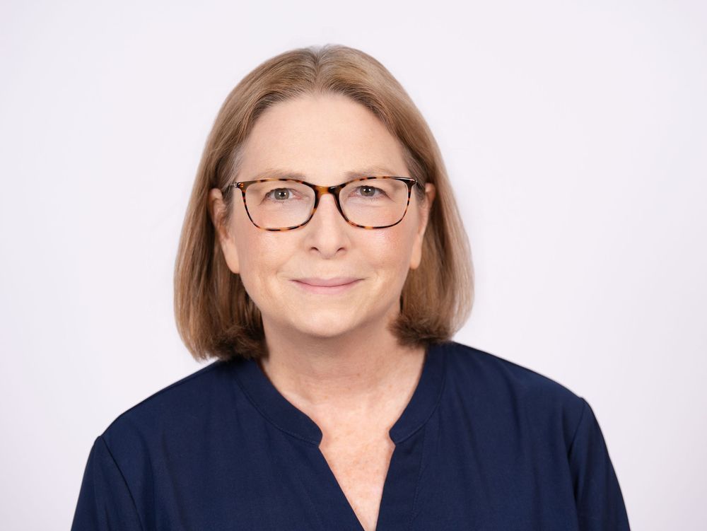 A woman with shoulder-length hair and tortoiseshell glasses in a navy blouse against a light backdrop