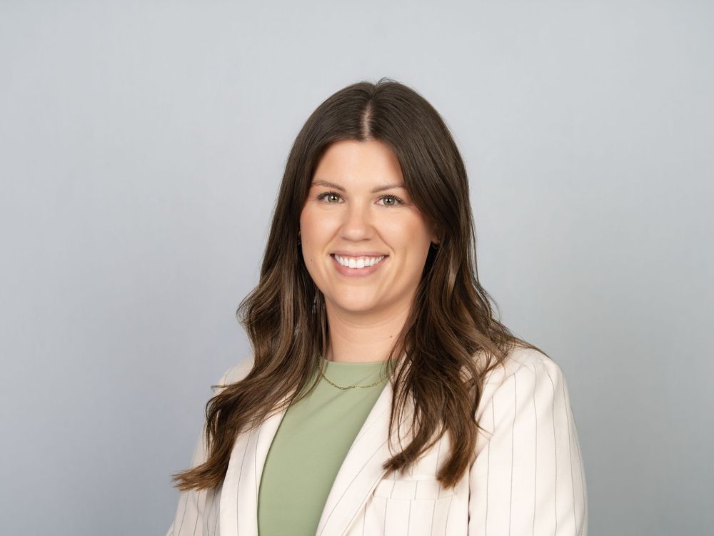 A smiling woman with long brown hair in a pinstripe blazer against a grey studio backdrop