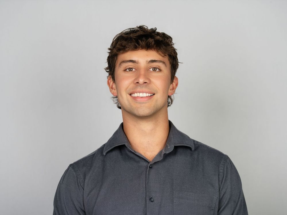 A smiling young man with curly hair in a charcoal button-up shirt against a grey backdrop