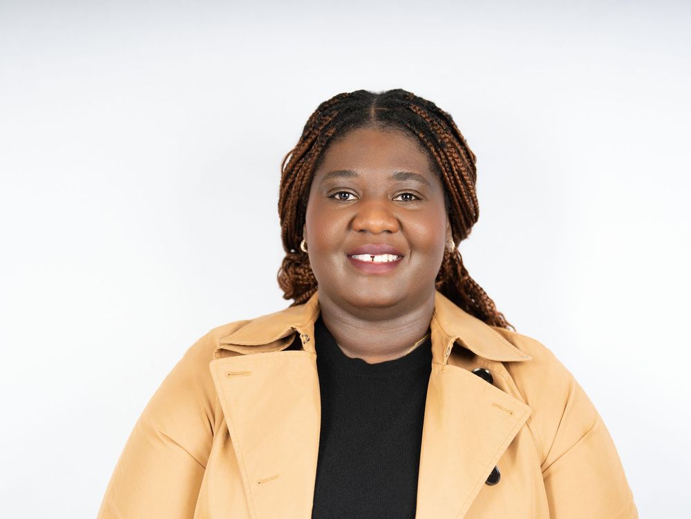 A smiling woman with long braids in a camel coat against a white studio backdrop
