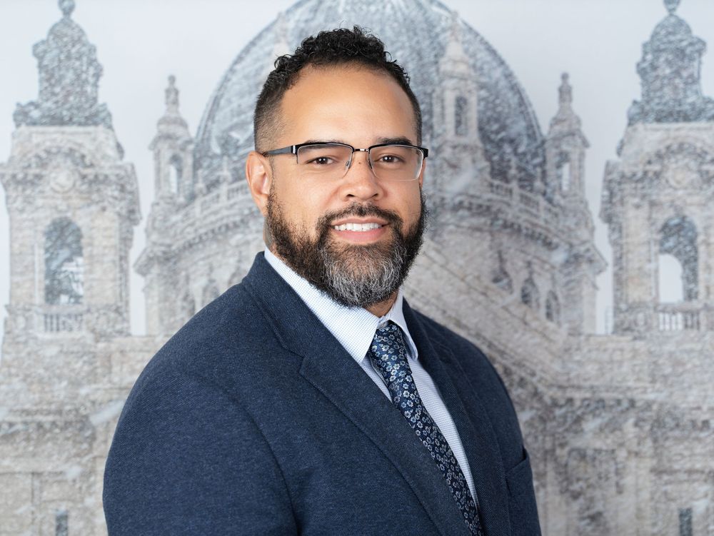 A smiling bearded man with glasses in a navy blazer and tie before a snowy domed cathedral