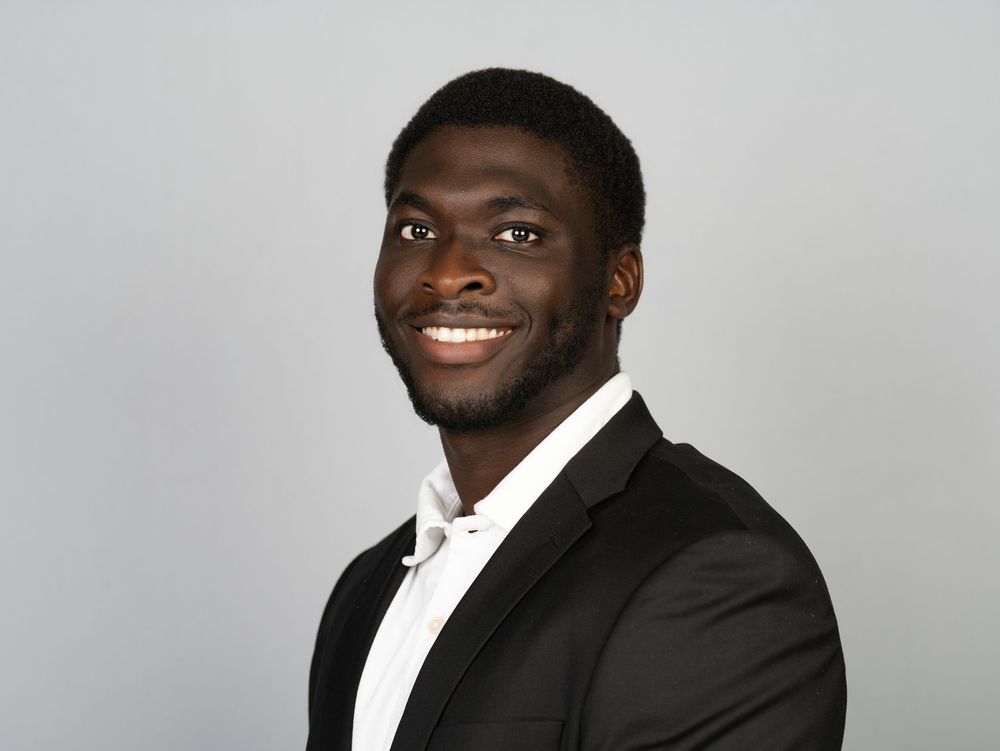A smiling young man in a black suit jacket and white shirt against a grey studio backdrop