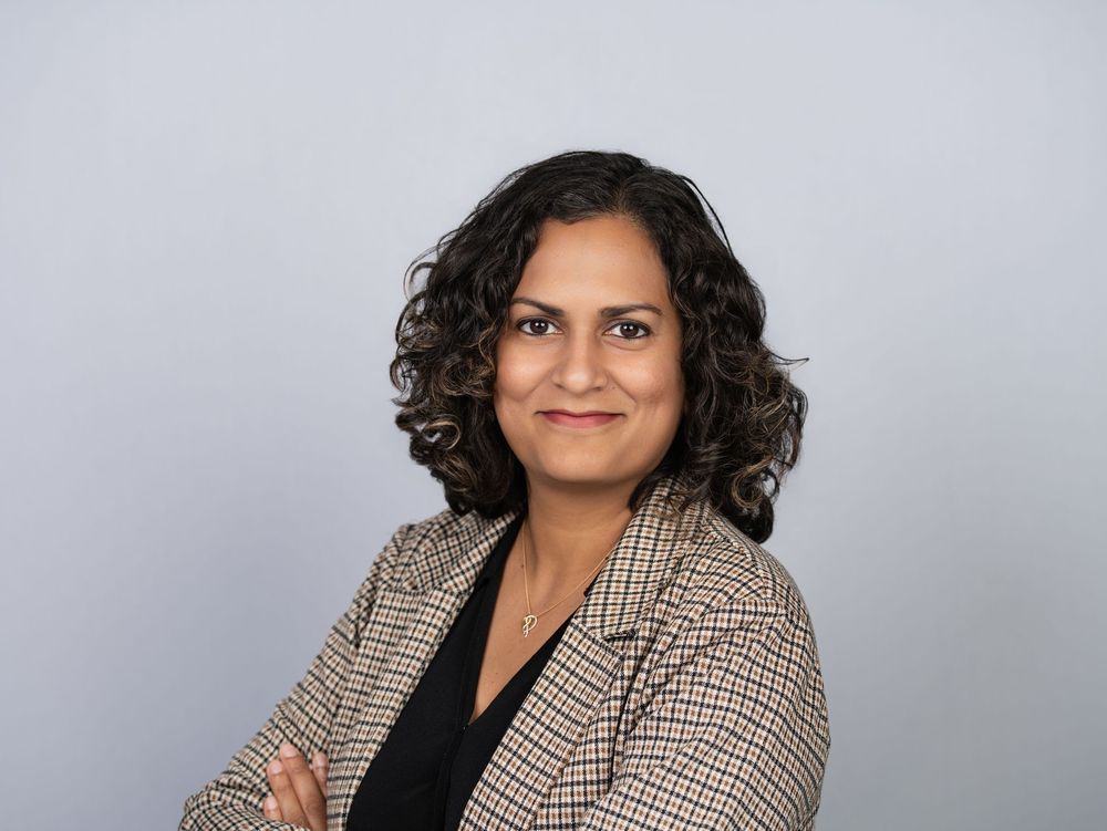 A woman with curly hair in a houndstooth blazer with arms crossed against a grey backdrop