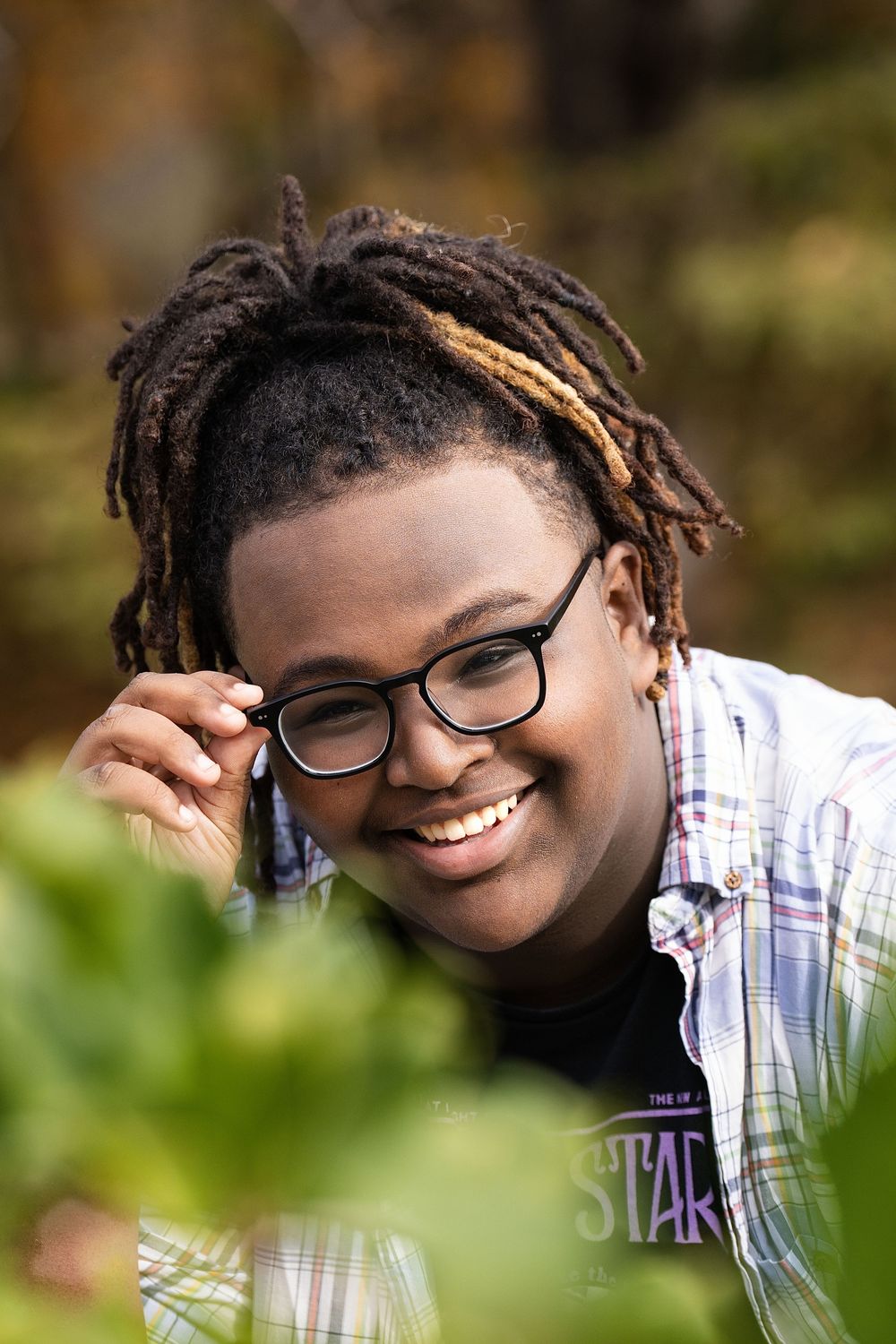 A smiling young person in glasses adjusting their frames while peeking through green leaves outdoors