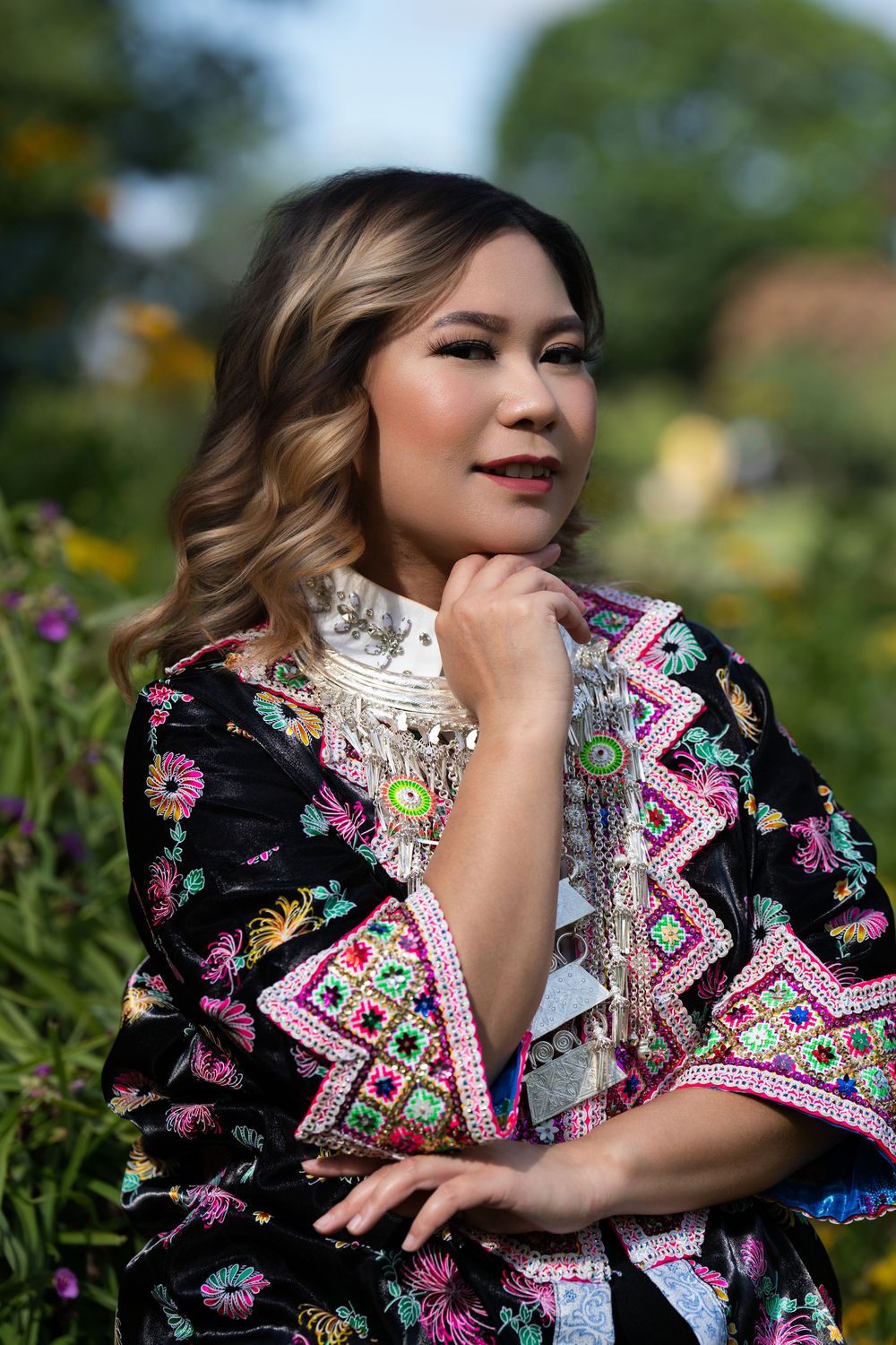 A woman in an embroidered jacket and silver collar necklace posing with hand to chin in a garden