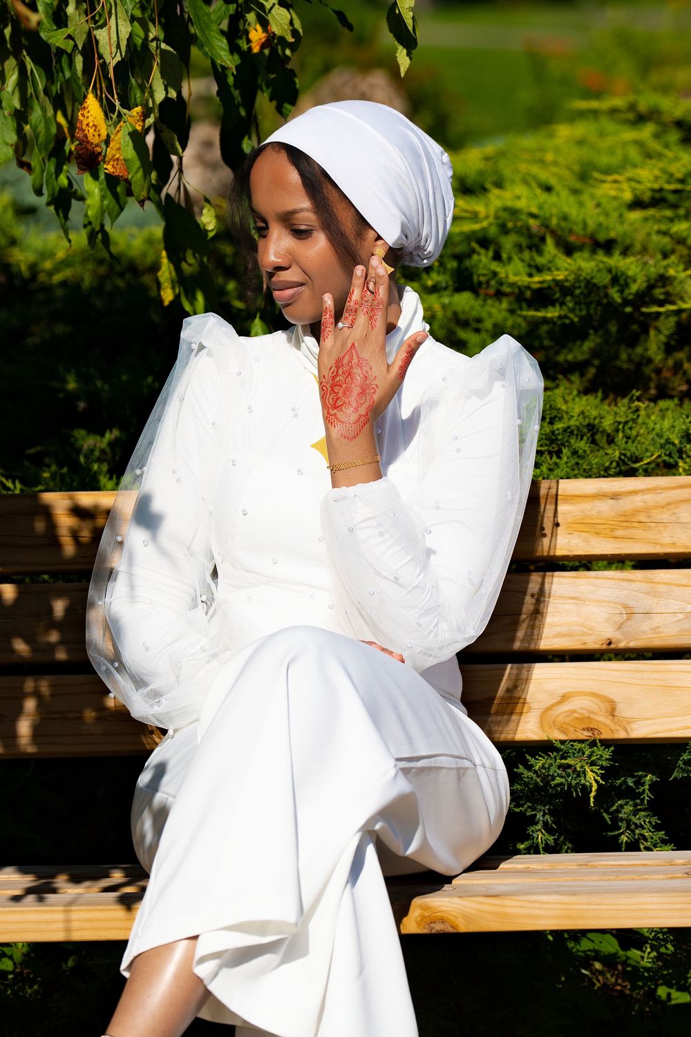 A woman in a white outfit and white head wrap sitting on a wooden bench among greenery