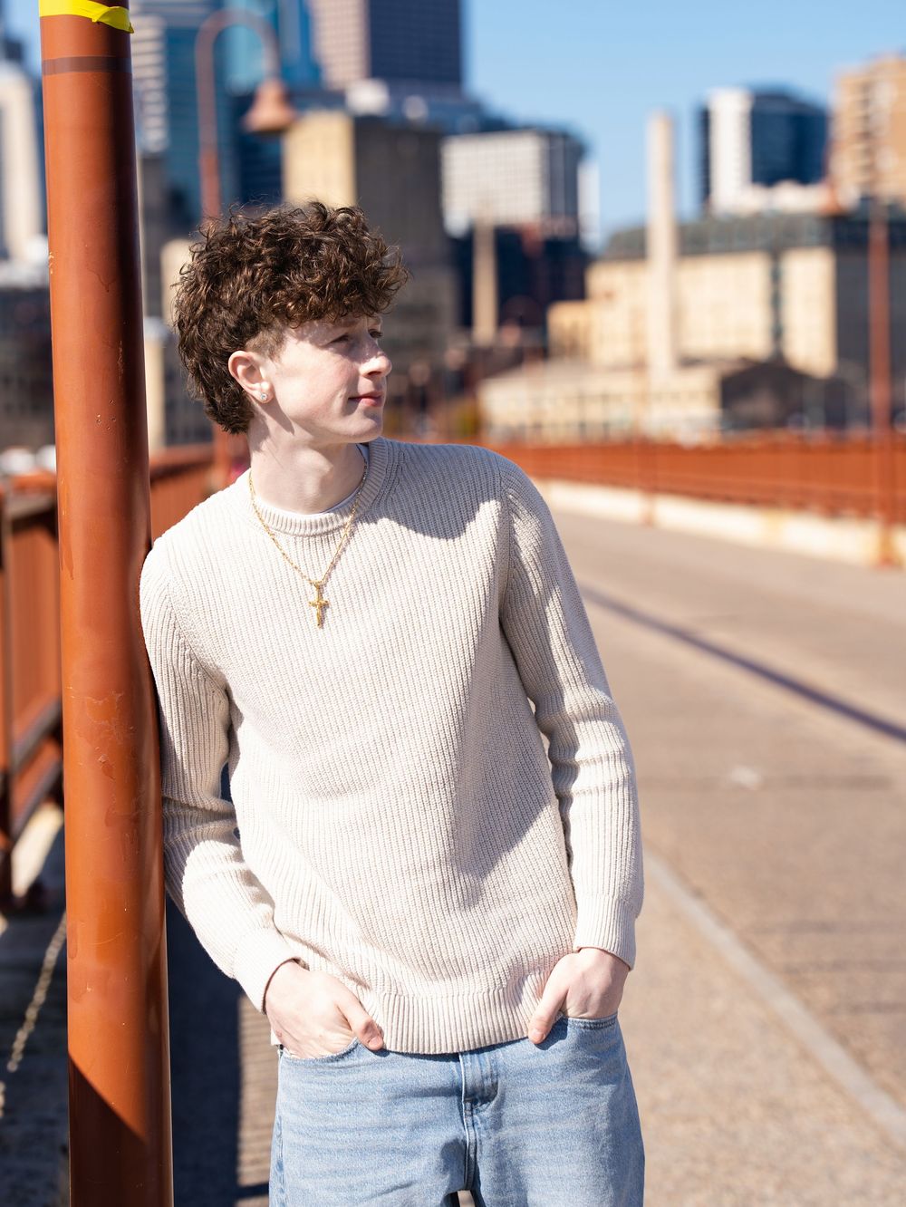 A young man in a cream sweater leaning on a bridge railing with a city skyline behind