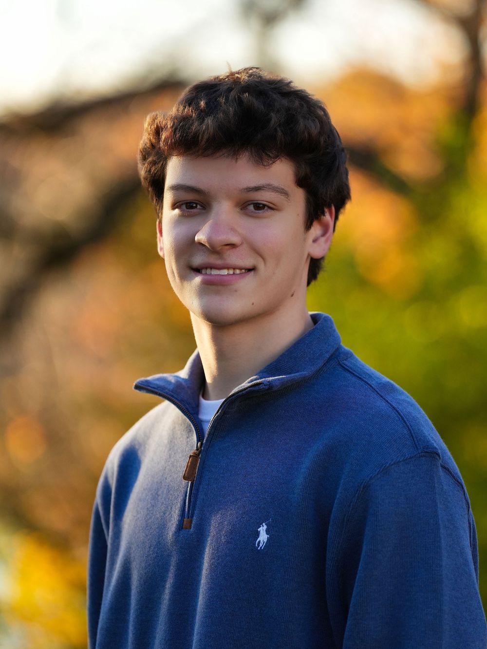 A smiling young man in a navy quarter-zip sweater outdoors among autumn trees