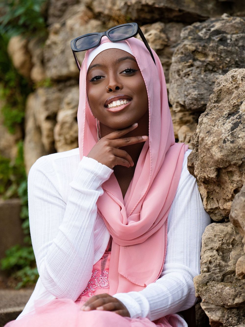 A smiling young woman in a pink headscarf and white cardigan seated beside a rock wall