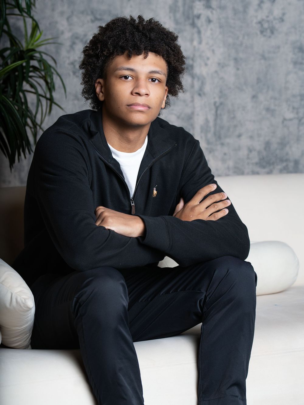 A young man in a black zip jacket sitting with arms crossed on a white studio sofa