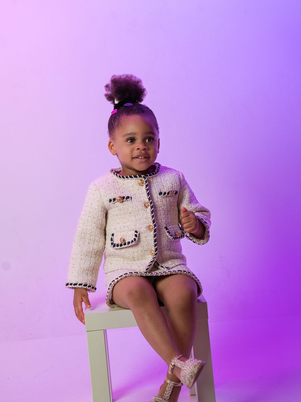A toddler in a tweed jacket and skirt sitting on a stool against a purple backdrop