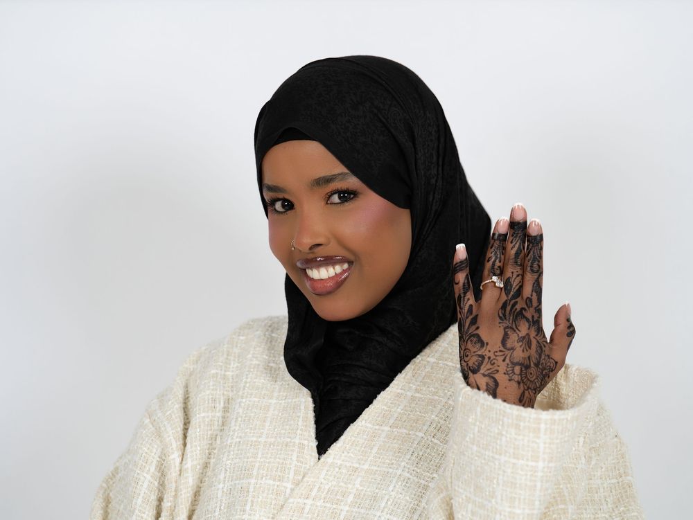 A smiling woman in a black headscarf raising a henna-patterned hand against a light studio backdrop