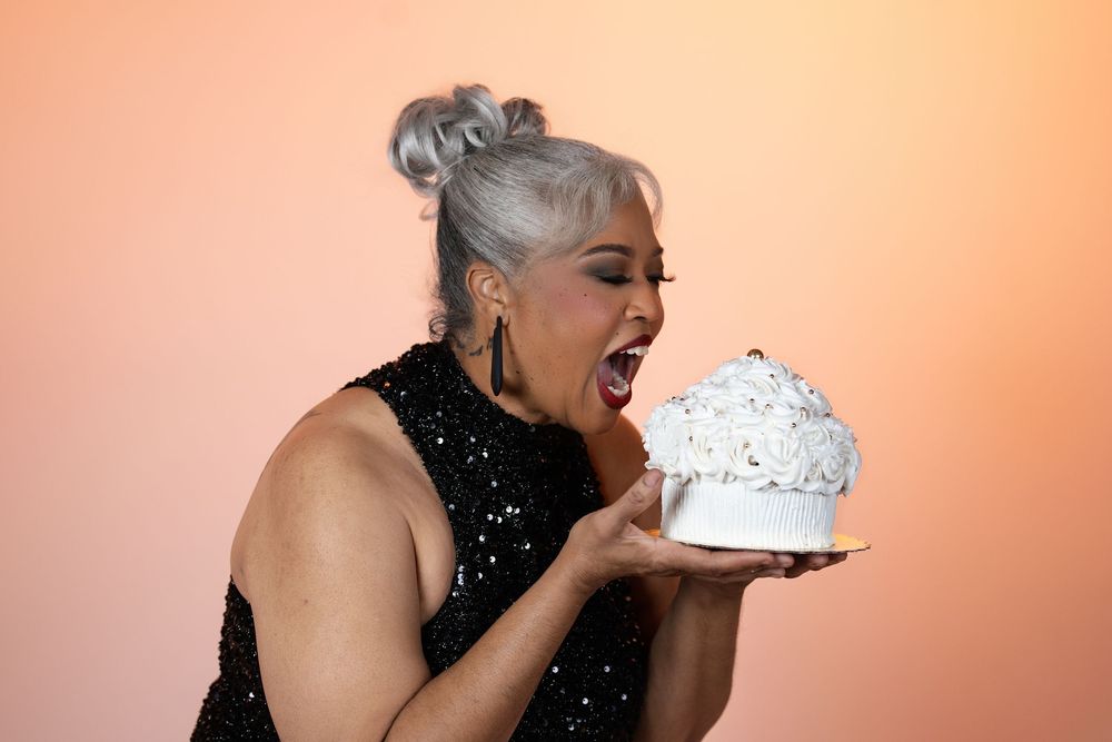 A woman with silver hair in a sequined top playfully biting a white cake, peach backdrop