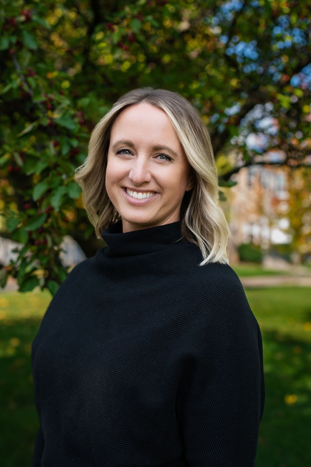 A smiling woman in a black turtleneck sweater standing in a leafy park