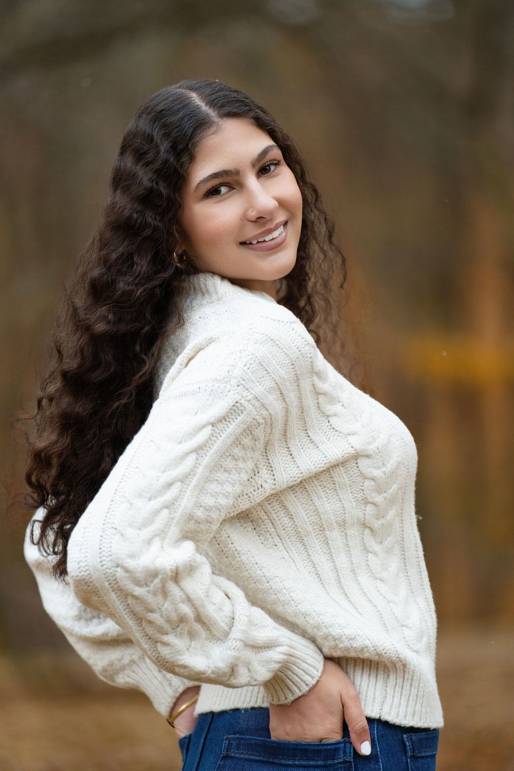 A young woman in a cream cable-knit sweater looking over her shoulder in autumn woods