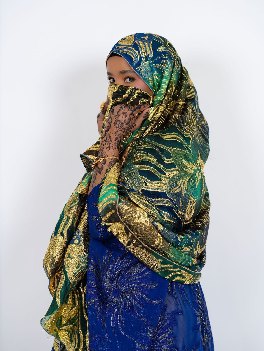 A woman in a green and gold patterned scarf drawn across her face against a white backdrop