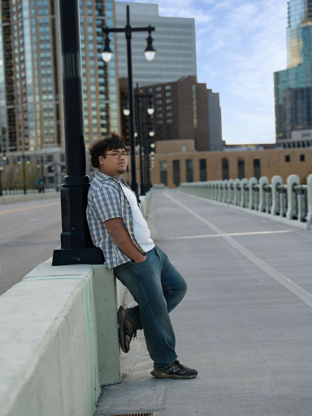 A young man in a plaid shirt leaning against a bridge wall with city buildings behind
