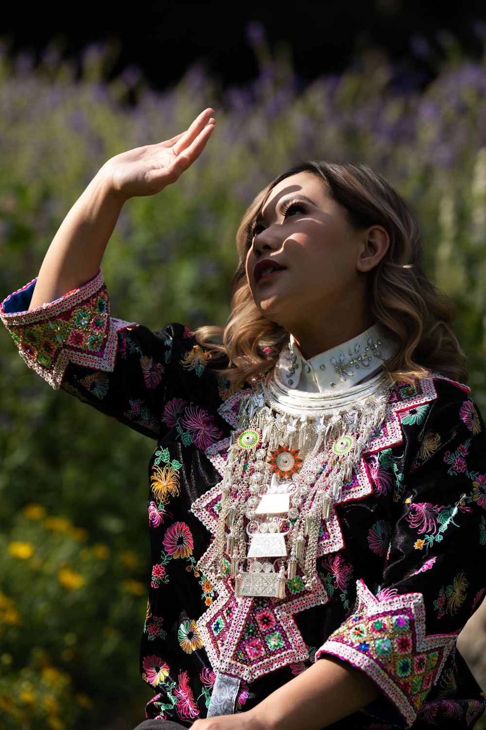 A woman in an embroidered jacket and silver necklace raising her hand toward the light among flowers