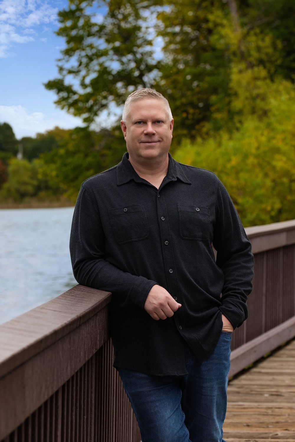 A man in a black shirt leaning on a wooden boardwalk railing beside a lake