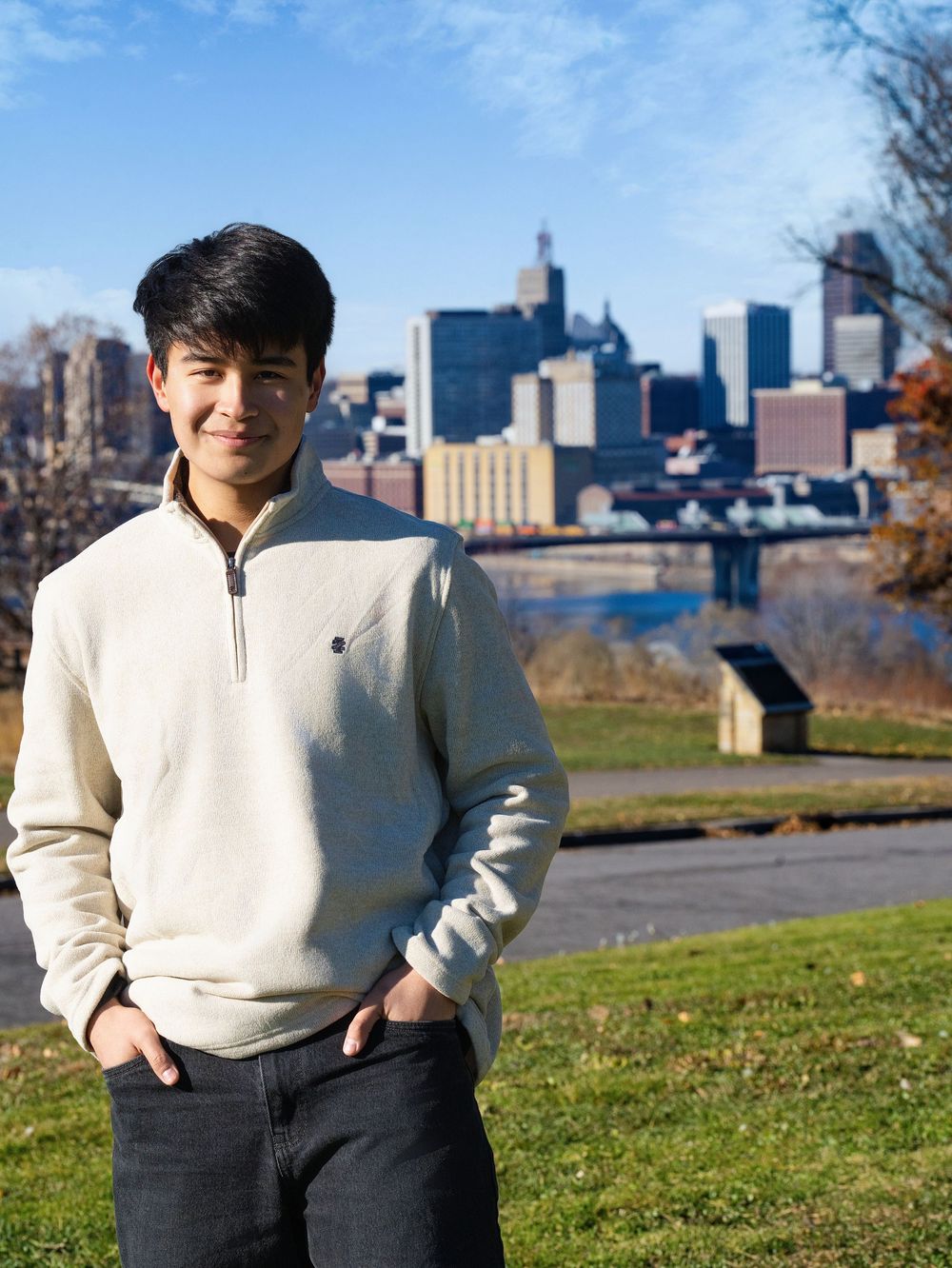 A young man in a cream quarter-zip sweater stands in a park overlooking a downtown skyline