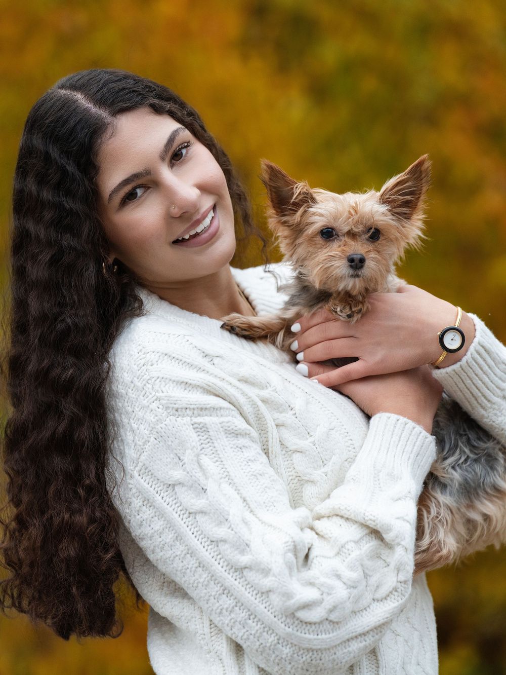 A young woman in a white cable-knit sweater holds a small terrier against autumn foliage