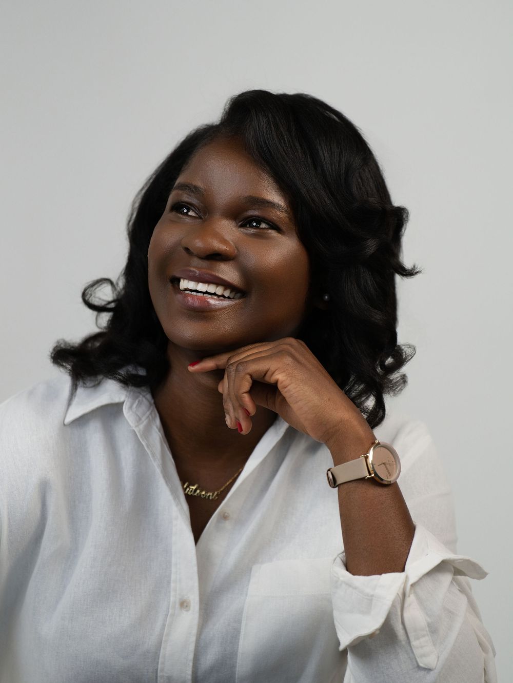 A woman in a white blouse smiles with her hand beneath her chin in a studio