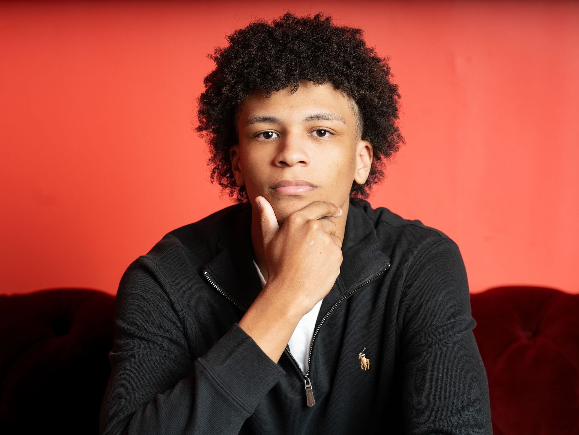A young man in a black quarter-zip rests his chin on his hand against a red wall