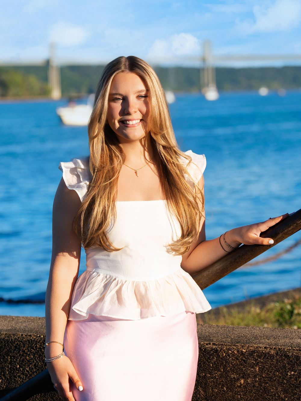 A young woman in a white top and pink skirt stands by a lake with sailboats