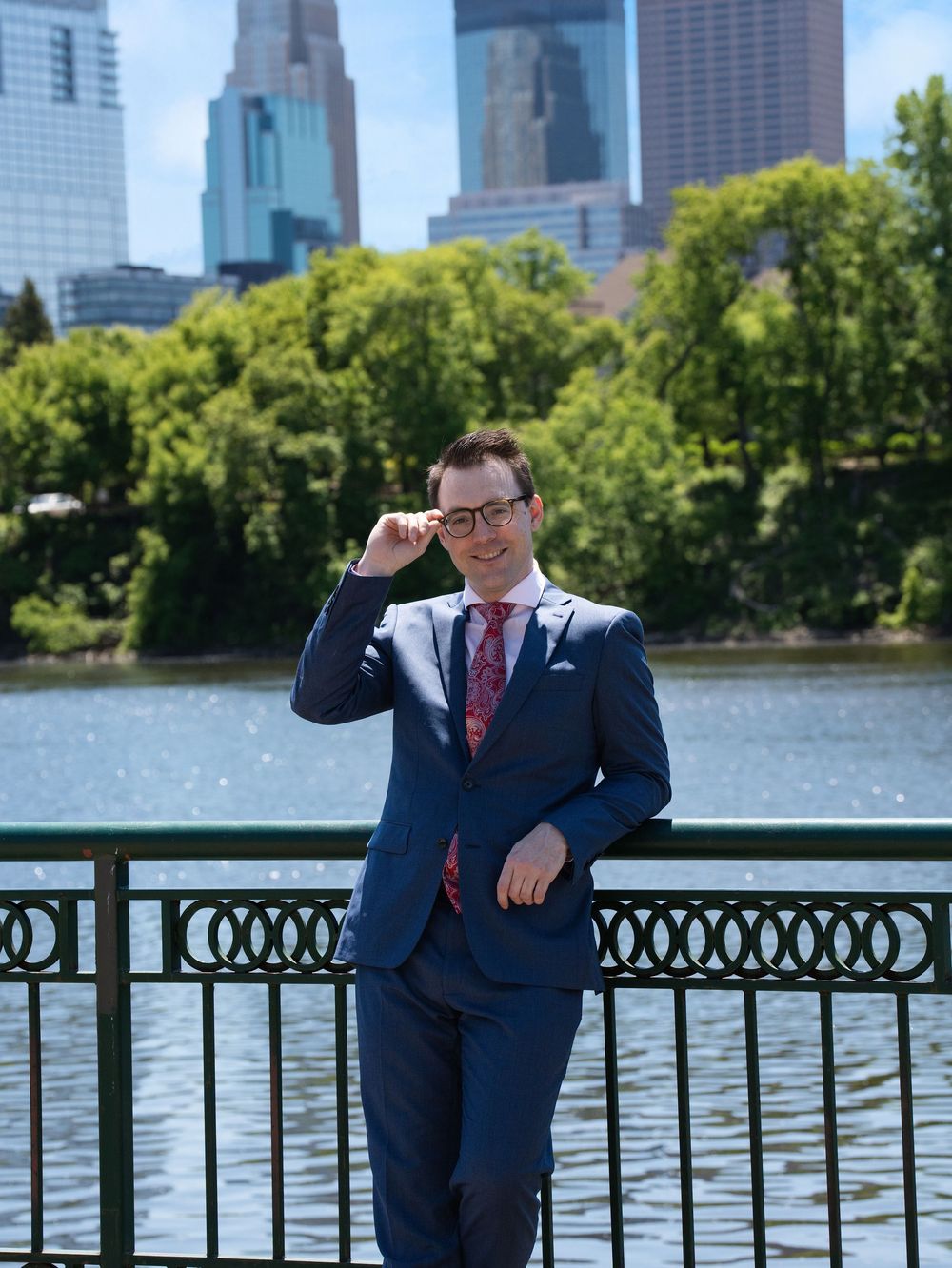 A man in a blue suit adjusts his glasses by a riverside railing below the Minneapolis skyline