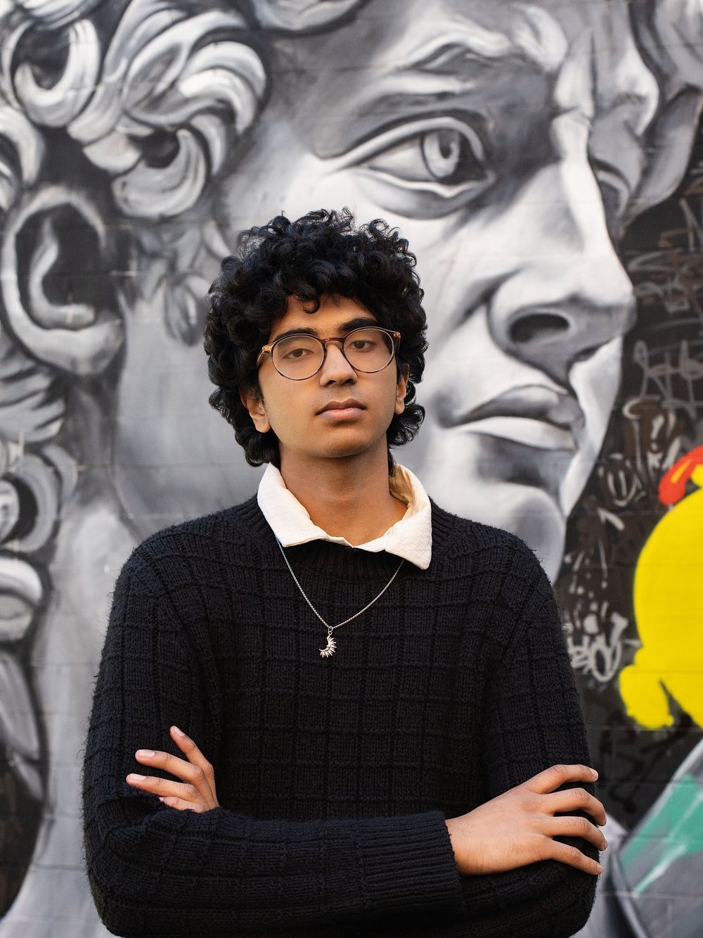 A young man in glasses stands with arms crossed before a black and white street mural