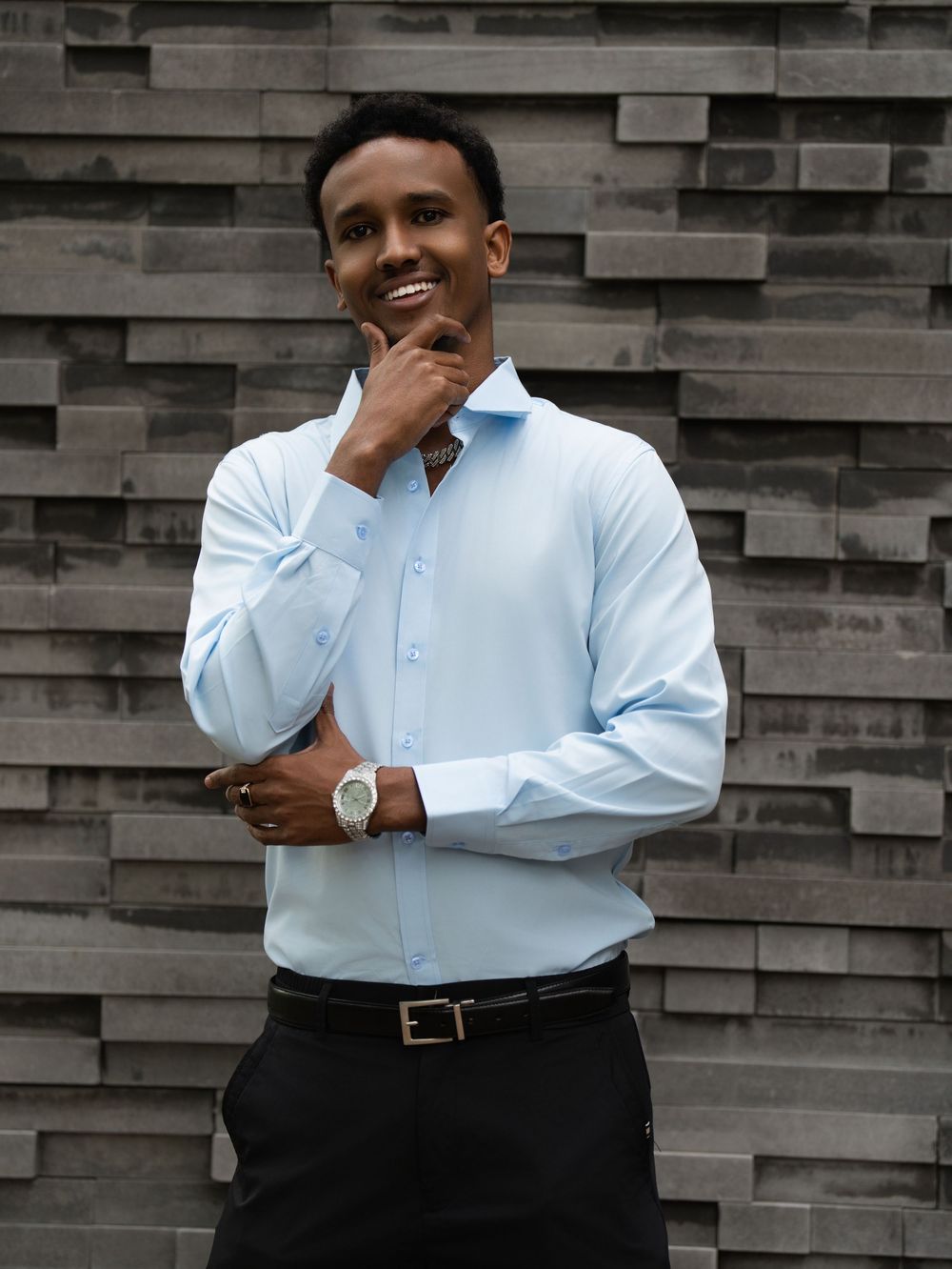 A young man in a light blue shirt poses with hand to chin against a stacked stone wall
