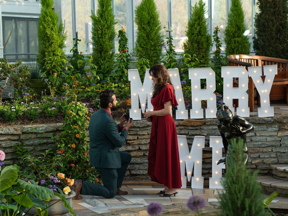 A man kneels to propose to a woman in red before lit MARRY ME letters in a garden