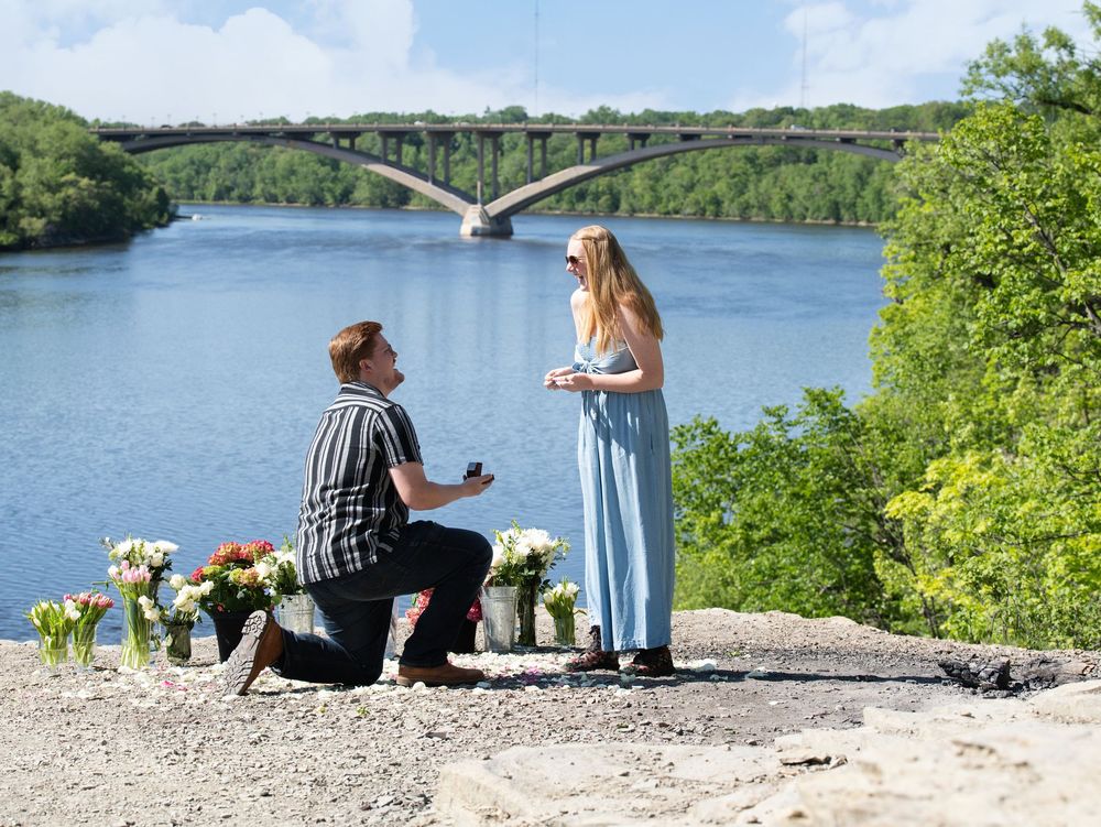 A man kneels to propose on a river bluff lined with flowers below an arched bridge