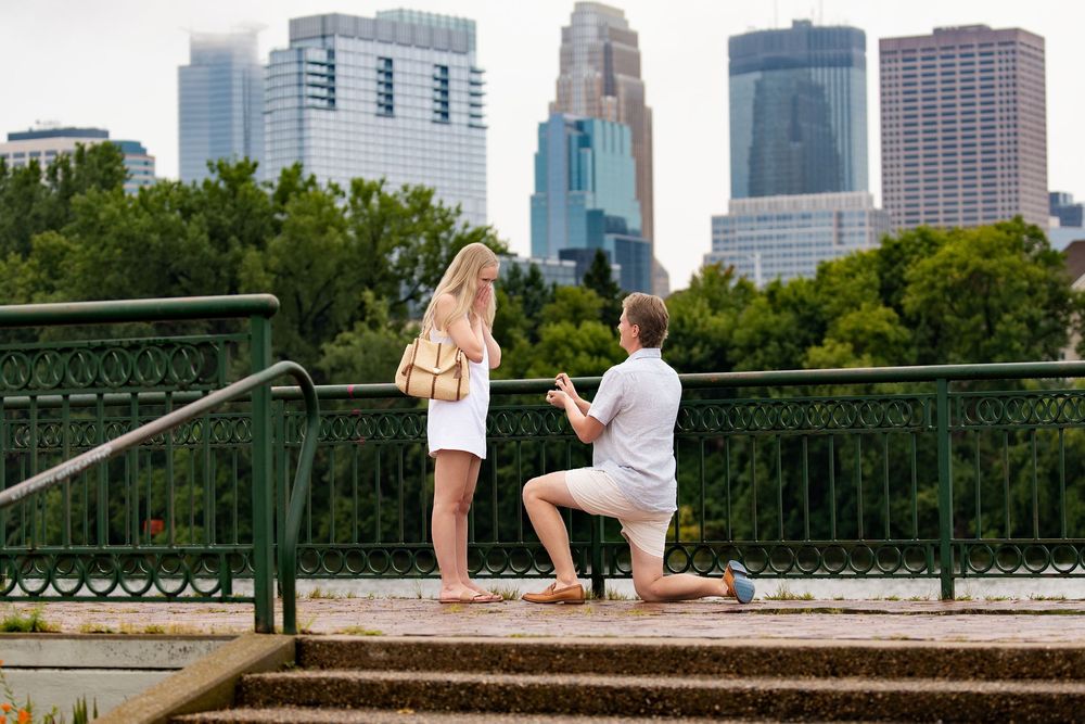 A man kneels to propose to a woman at a railed overlook below the Minneapolis skyline