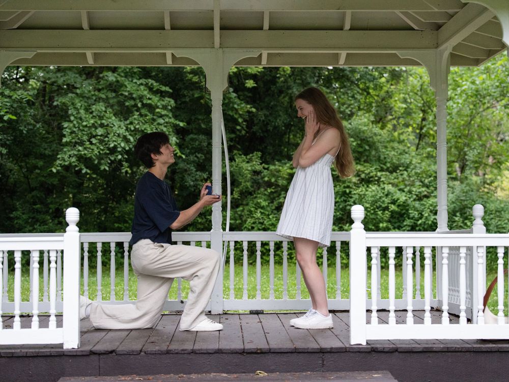 A man kneels to propose to a woman in a white sundress inside a white garden gazebo