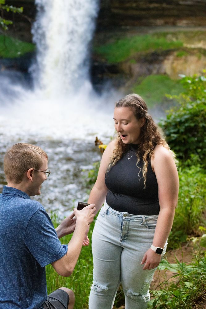 A man kneels to propose to a surprised woman in front of a rushing waterfall