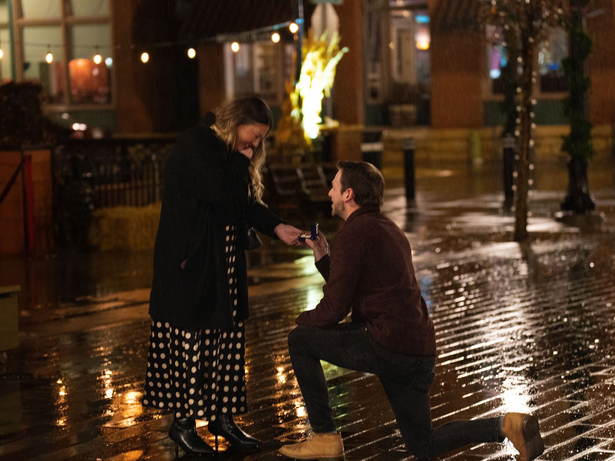 A man kneels to propose to a woman on a rain-slicked street at night