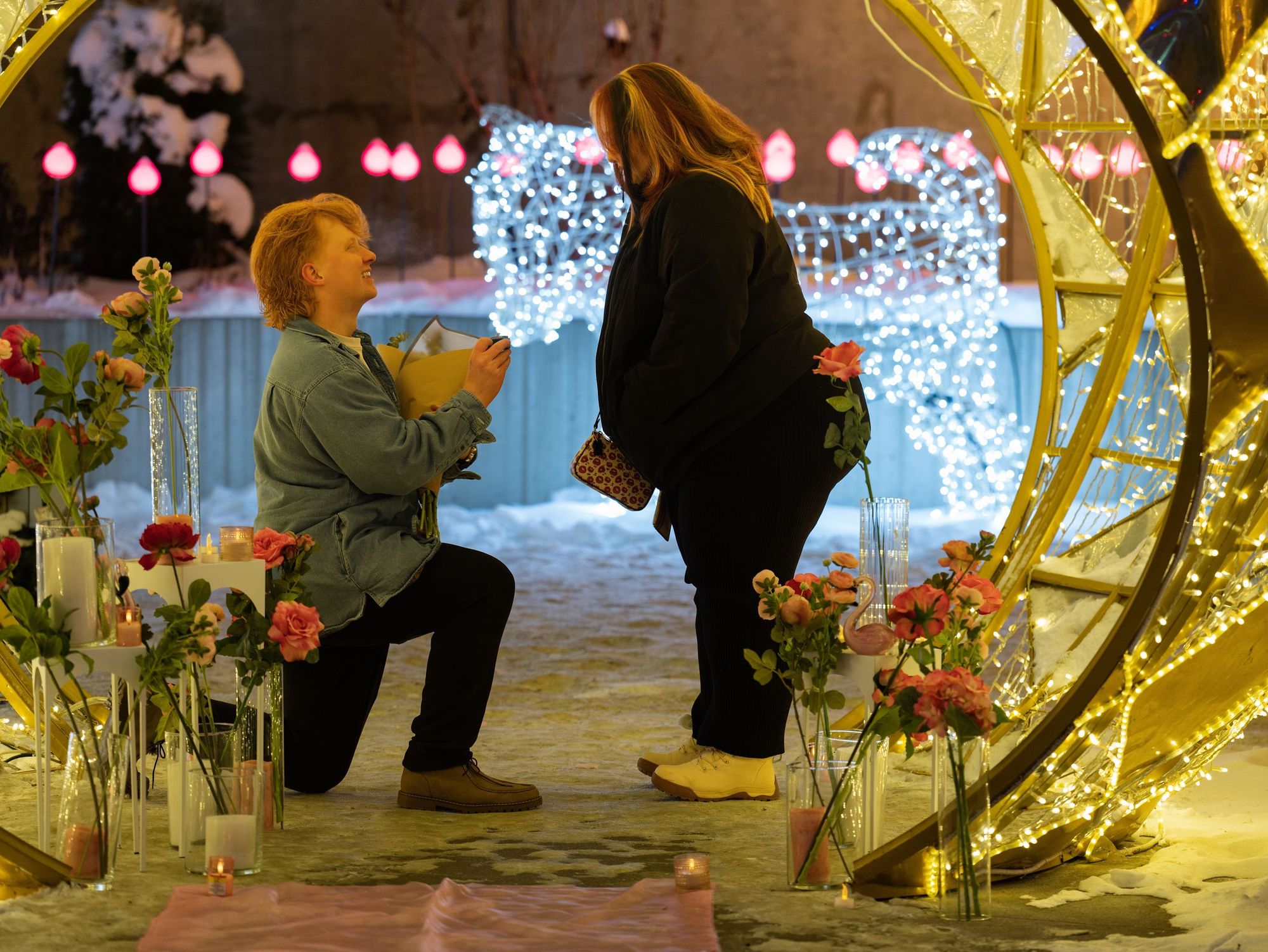 One partner kneels to propose among roses, candles and glowing light sculptures on a snowy night